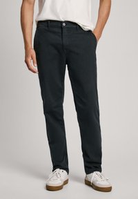 Tumed puuvillasegusega chinos, millel on eestaskud, nupuga sulgemine ja sirge jala kujundus, koos valgete tossudega.