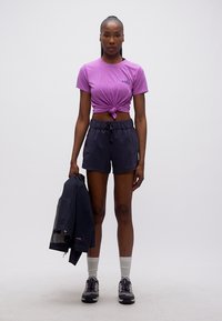 Γυναίκα που στέκεται με το σώμα στραμμένο προς τα εμπρός, φοράει μωβ μπλούζα με κόμπο, μαύρο σορτς, κρατάει ένα μαύρο μπουφάν στο ένα χέρι, λευκές κάλτσες και αθλητικά παπούτσια.