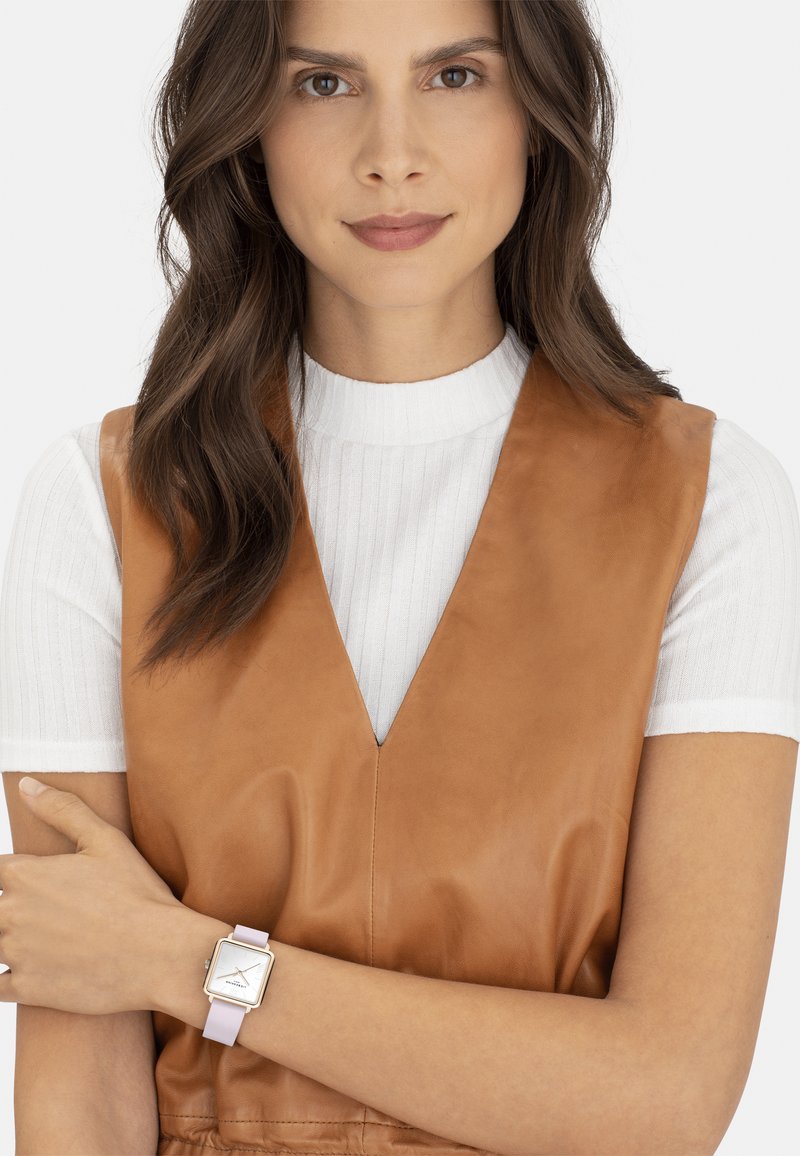 Bruine leren vest over een witte geribbelde top. Vierkante horloge met een lavendelkleurige band en zilveren accenten om de pols. Zachte texturen en strakke lijnen.