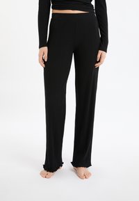 Vrouw in zwarte grofgebreide wijde broek met gerimpelde zoom, staand op blote voeten voor een witte achtergrond, met ontspannen handen langs haar zij.