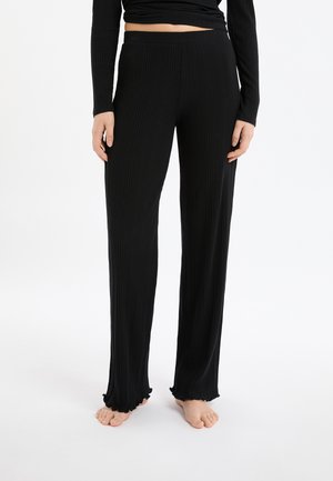 Kobieta w czarnych szerokich spodniach z prążkowanego materiału z falbankami na krawędziach, stoi boso na białym tle, ręce swobodnie wzdłuż ciała.