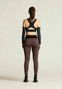 Schwarzer Sport-BH mit offener Rückseite, kombiniert mit braunen Leggings. Beinhaltet schwarze Armschoner und schwarze Turnschuhe. Glatte Textur, körperbetontes Design.