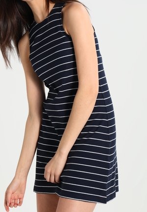 Mujer que lleva un vestido azul marino sin mangas con rayas horizontales blancas, de pie frente a un fondo claro y liso.