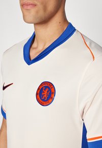 Vit fotbollströja med blå och orange accenter, med en blå v-ringning och en rund Chelsea FC-logga på bröstet.