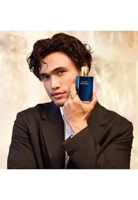 Davidoff Cool Water Duft in einer blauen Glasflasche mit goldenem Deckel. Ein Modell in einem schwarzen Anzug hält die Flasche vor einem verschwommenen Hintergrund.