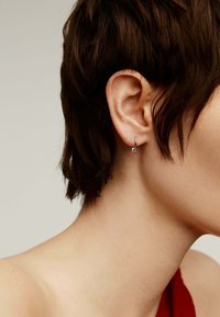 Pendiente de aro de plata con un diseño retorcido, llevado en una oreja de cabello corto. Superficie suave y acabado pulido. Tono de piel parcialmente visible.