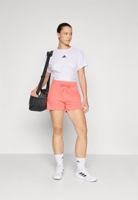 T-shirt atlético branco com logotipo preto, calções corais com cordão, e ténis brancos. Bolsa preta com alça para o ombro. Fundo branco.