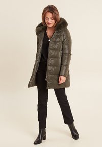 Gevoerde olijfgroene jas met een met bont gevoerde capuchon, drukknoopsluitingen en verticale stiksels. Draag deze over een zwarte top en broek, gecombineerd met zwarte laarzen met hak.