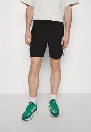 Zwarte cargoshorts met een elastische tailleband, voorzien van zijzakken en een losse pasvorm, gecombineerd met groene sneakers en witte sokken.