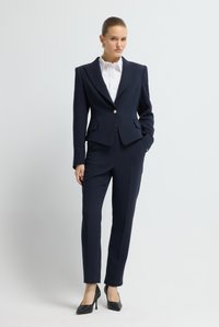 Marynarka w kolorze granatowym o dopasowanym kroju, ze wzmocnionymi ramionami, pojedynczym złotym guzikiem z przodu oraz dopasowanymi spodniami. Noszona z białą koszulą z kołnierzykiem i czarnymi szpilkami.