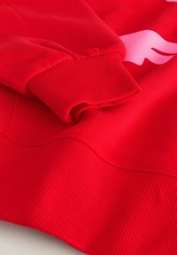 Hanorac roșu cu manșete și tiv cu nervuri, textură netedă și un design grafic mare de culoare roz pe față.