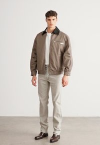 Brūna bumbvedēja jaka ar melnu apkakli, priekšpusē ar rāvējslēdzēja kabatu. Valkāta virs baltas T-krekls un gaiši pelēkiem džinsiem, kombinēta ar melniem apaviem.