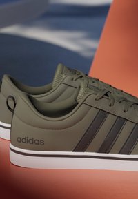 Par de zapatillas Adidas verde oliva con rayas negras y suelas blancas colocadas sobre una superficie naranja con un fondo azul desenfocado.