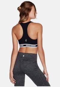 Sostén deportivo negro con diseño de espalda nadadora, que cuenta con una banda de marca en blanco. Llevado con leggings de tiro alto en un gris oscuro texturizado.
