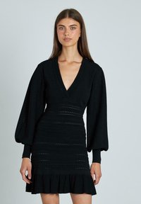Abito nero lavorato a maglia con scollatura a V profonda, maniche lunghe a sbuffo, motivi testurizzati e orlo a volant. Presenta una silhouette aderente.