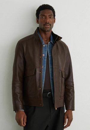 Mann trägt geschichtete braune Leder- und blaue Denimjacken, steht vor einer schlichten weißen strukturierten Wand.
