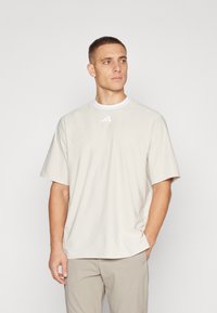 Tricou sportiv bej deschis, cu guler rotund, mâneci scurte și un logo alb subtil pe piept. Material neted și croială lejeră.