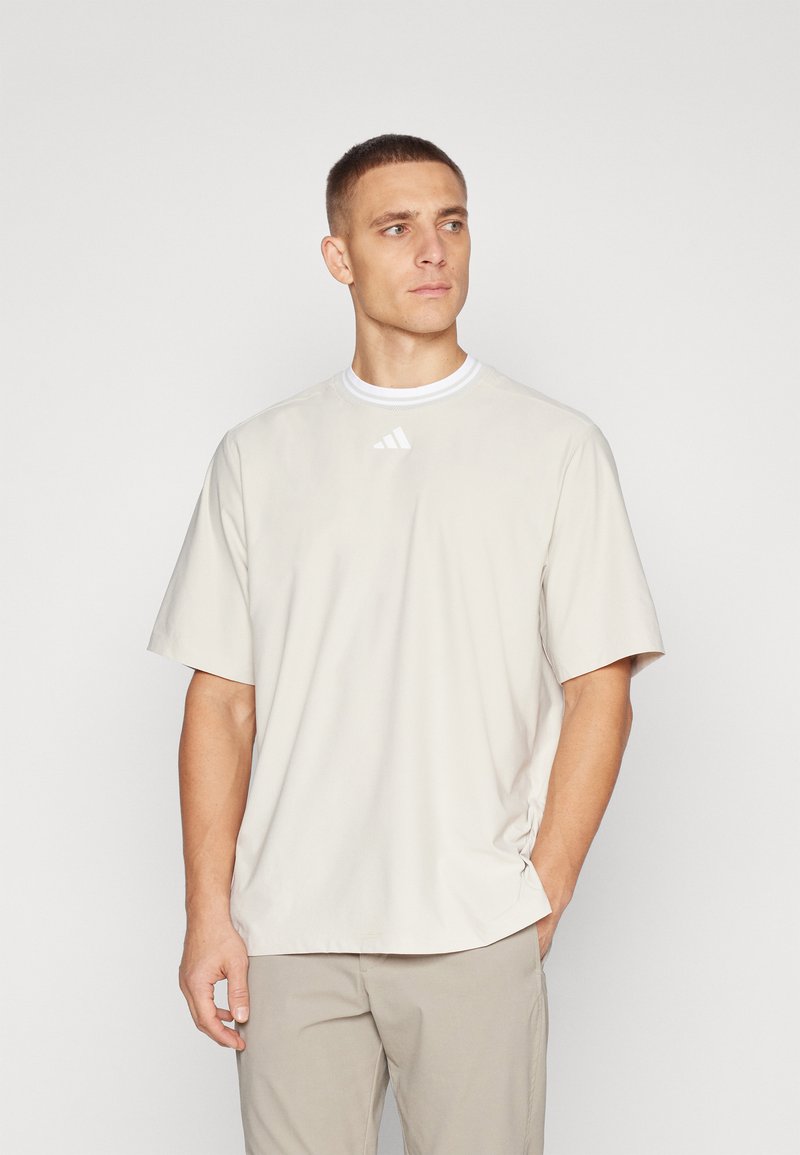 Tricou sportiv bej deschis, cu guler rotund, mâneci scurte și un logo alb subtil pe piept. Material neted și croială lejeră.
