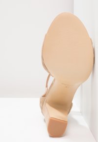 Escarpin en daim nude à talon haut avec un bout arrondi lisse, une semelle texturée et une bride de cheville. Marqué de la taille 37 au dessous.