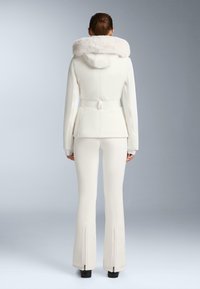 Giacca bianca con cappuccio in pelliccia sintetica, vestibilità sagomata, vita aderente e pantaloni coordinati aderenti con cerniere alle caviglie. Texture liscia complessivamente.