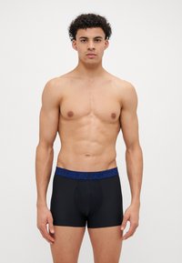 Crne bokserice s plavim logotipom "UNDER ARMOUR" na pojasu, s prilagođenim dizajnom i mekim, rastezljivim materijalom.