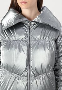 Casaco puffer prateado com gola alta, textura brilhante, costura horizontal e um fecho frontal com puxador circular.