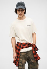 Νέος άνδρας που φοράει μπεζ μπλουζάκι McLaren, κόκκινο και μαύρο καρό πουκάμισο δεμένο γύρω από τη μέση, παντελόνι καμουφλάζ cargo και σκούρο γκρι καπέλο.