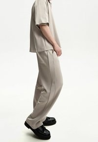 Beige top met korte mouwen en bijpassende broek van een glad, zacht materiaal. De broek heeft zijnaden en zakken. Zwarte schoenen maken de outfit compleet.