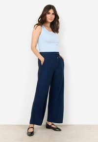 Lichtblauw tanktop en marineblauwe broek met wijde pijpen en zakken. De broek heeft een tailleband met trekkoord en een zachte, gestructureerde stof. Zwarte schoenen.