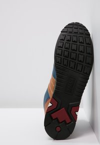 Sneakersula med en texturerad svart gummiyta, med en röd och grå geometrisk design och märkeslogotyp vid hälen.