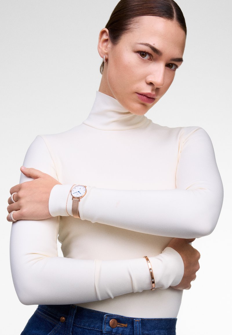 Top blanco de manga larga con cuello alto y un diseño sin costuras. Reloj de oro rosa con pulsera de malla y un brazalete a juego en la muñeca.