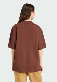 Camiseta oversized marrón de manga corta, hecha de tela suave. Presenta un diseño sencillo y un ajuste relajado, combinada con pantalones beige.