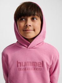 Kind met bruin haar dat een roze Hummel sporthoodie draagt, kijkt iets naar rechts met een neutrale uitdrukking tegen een effen achtergrond.