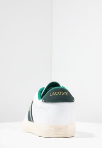 Bílé tenisky Lacoste se zelenými akcenty a logem; svrchní materiál z texturované kůže; krémová gumová podrážka; zadní část seproměněnými zlatými písmeny a polstrovaným límcem.