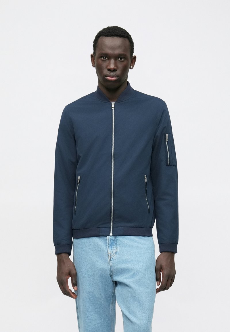 Junger Mann, der eine navyblaue Bomberjacke mit Reißverschlüssen an der Vorderseite und den Ärmeln sowie hellblauen Jeans trägt, vor einem schlichten weißen Hintergrund steht.