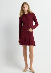 Bordeaux ritsjack met een gestructureerd patroon, gecombineerd met een bijpassende geplooide rok met twee voorkepers. Beige schoenen maken de look compleet.