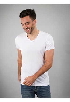 Lächelnder Mann, der ein schlichtes weißes V-Ausschnitt-T-Shirt und blaue Jeans trägt, vor einem grauen Hintergrund stehend, mit einer leicht erhobenen Hand.
