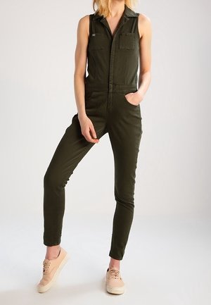 Kombinezon bez rękawów w oliwkowej zieleni z dwoma przednimi kieszeniami, dopasowany fason i zwężane nogawki, zestawiony z jasnobrązowymi tenisówkami.