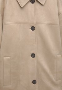 Beige faux suede jasje met een kraag, voorzien van vijf donkere knopen, twee zijzakken en gestikte details over de borst.
