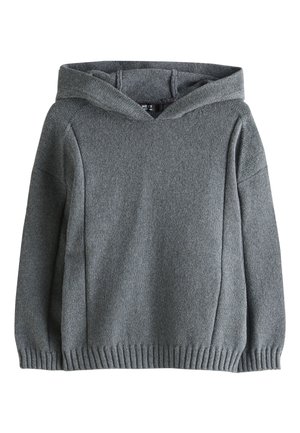 Junge mit lockigem Haar, der einen grauen Kapuzenpullover, schwarze Cargohosen und weiße Turnschuhe trägt, steht vor einem einfarbigen beigen Hintergrund.