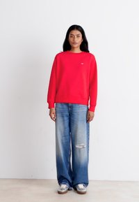 Red cotton sweatshirt with a crew neckline, paired with light blue distressed wide-leg jeans and metallic slip-on shoes. Simple design.