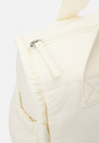 Sac rembourré couleur crème avec fermeture éclair et tissu texturé. Il possède une sangle beige et une couture décorative sur le côté.