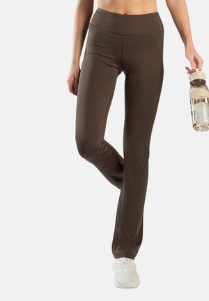 Person trägt braune, eng anliegende Hose und weiße Sneakers, steht mit einem angewinkelten Bein und hält eine klare Wasserflasche mit beigem Griff.