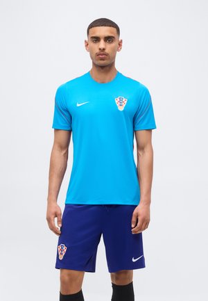 Mężczyzna w niebieskiej koszulce bramkarza reprezentacji Chorwacji i ciemnoniebieskich spodenkach z logo Nike, stojący na jednolitym jasnym tle.