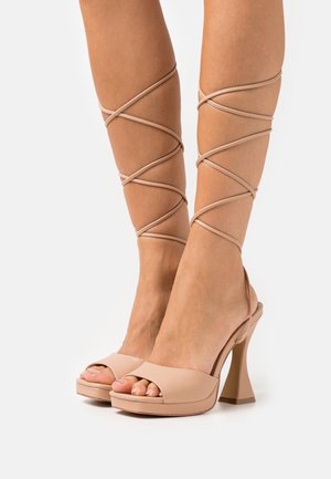 Chaussures à talons hauts beige avec orteils ouverts, dotées d'un talon épais et de brides croisées autour des chevilles. Matériau lisse avec une finition brillante.