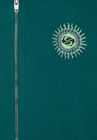 Giacca zip-up turchese con un logo a raggi di sole in verde e nero sul lato sinistro del petto, realizzata in un tessuto morbido con una texture liscia.