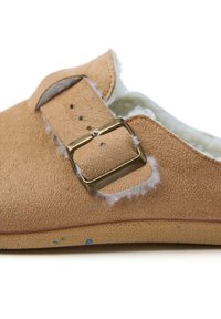 Chausson en suede brun clair avec un intérieur doux, doté d'une boucle dorée et de coutures décoratives. Semelle plate avec des détails texturés.