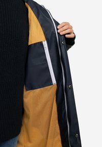 Námořnická voděodolná bunda se žlutou síťovinou, která má zipovou kapsu a patentní knoflíky vpředu.