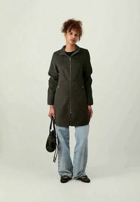 Μακριά, σκούρα γκρι παλτό με φερμουάρ μπροστά, δύο μπροστινές τσέπες και γιακά. Συνδυάζεται με ανοιχτό μπλε παντελόνι με φαρδιά μπατζάκια και μαύρα επίπεδα παπούτσια.