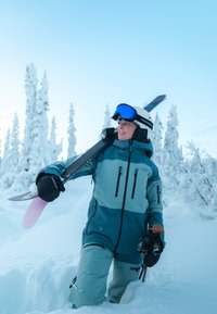 Skitøj omfatter en vandtæt jakke i blågrøn og grå med lynlåslommer, matchende bukser, sorte handsker og en hvid hjelm med blå goggles.